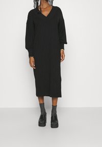 Robe noire en tricot avec un col en V, manches longues bouffantes, texture côtelée et longueur midi. Portée avec des chaussures à plateforme foncées et des chaussettes à motifs.