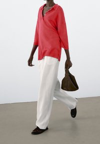 Koralowa dzianinowa koszulka polo na trzy guziki, zestawiona z luźnymi białymi spodniami i ciemnooliwkową zamszową torebką. Całość dopełniają czarne sandały.