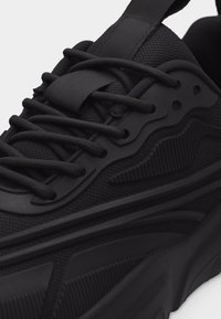 Μαύρο αθλητικό παπούτσι με υφασμάτινο πλέγμα, χοντρή σόλα, μαύρα κορδόνια και τονικές συνθετικές επενδύσεις σε κοντινή λήψη.