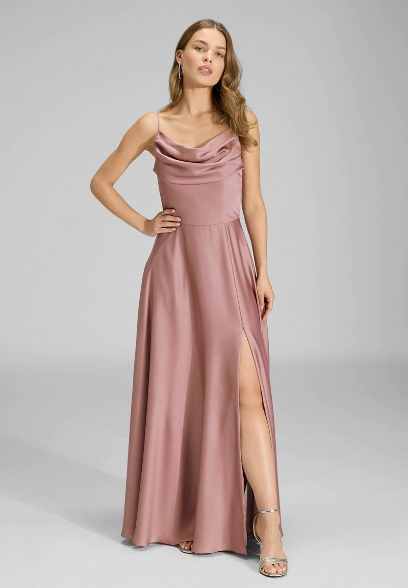 Femme en robe satinée mauve à encolure drapée et fente haute, portant des talons argentés à fines lanières, posant avec une main sur la hanche devant un fond gris.