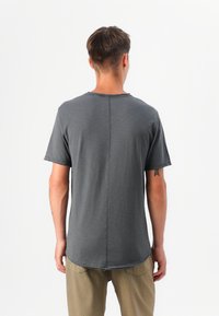 Modelo masculino visto por la espalda, con una camisa de manga corta gris y pantalones beige, contra un fondo blanco liso.