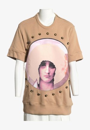 Chemise à manches courtes couleur fauve avec un grand imprimé circulaire d'un visage féminin stylisé et des clous en laiton autour du cercle.