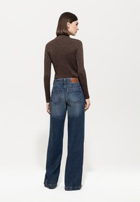 Brauner, glitzernder Rollkragenpullover kombiniert mit weit geschnittenen Denim-Jeans, die einen Gradienten von hell zu dunkel aufweisen und ein braunes Lederpatch auf der Rückseite haben.