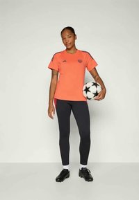 Ryškiai oranžinė sportinė marškinė su juodomis juostelėmis, derinamos su juodomis leggings. Modelis laiko juodai baltą futbolo kamuolį, avėdamas sportbačiais.