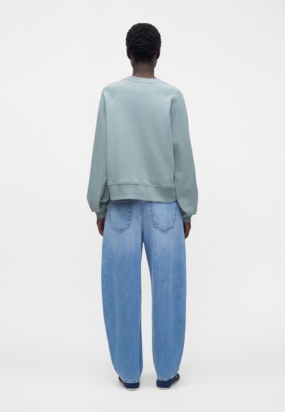 Personne aux cheveux courts et bouclés portant un sweat-shirt vert clair et un jean bleu clair ample, debout de dos sur un fond uni.