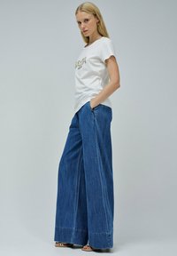 Camiseta blanca de manga corta con letras doradas brillantes, combinada con unos vaqueros de pierna ancha en azul, que cuentan con bolsillos laterales y un dobladillo limpio.