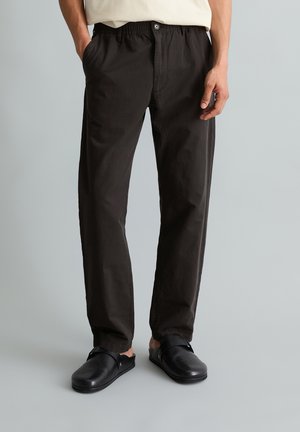 Man wearing black straight-leg pants with elastic waistband and black slip-on shoes, standing with hands in pockets against gray background.