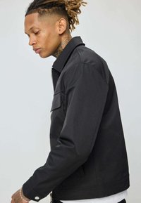 Jeune homme avec des tatouages et des dreadlocks portant une veste noire zippée par-dessus une chemise blanche, tourné vers la gauche contre un fond uni.