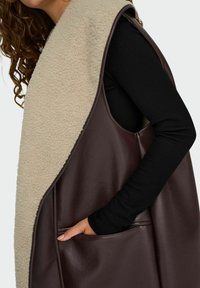 Brązowa skórzana kamizelka z beżowym kołnierzem z polarowego materiału, z boczną kieszenią i eleganckim krojem, podkreślająca gładkie materiały i kontrastujące faktury.