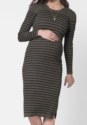 Zwangere vrouw die een olijfgroen geribd jurkje met lange mouwen en zwarte horizontale strepen draagt, en haar buik vasthoudt met één hand.
