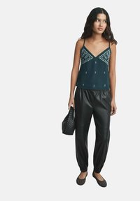 Top senza maniche verde scuro con accenti ricamati, abbinato a pantaloni jogger in ecopelle nera. L'outfit include scarpe basse marroni e una borsa testurizzata.