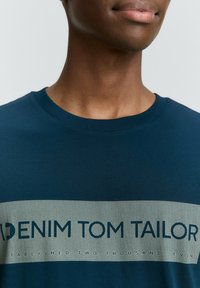 Jūras zilas kokvilnas t-krekls ar apaļu kakla izgriezumu. Piemīt izteikta gaiši pelēka horizontāla josla ar tekstu "DENIM TOM TAILOR" un dibināšanas datumu.