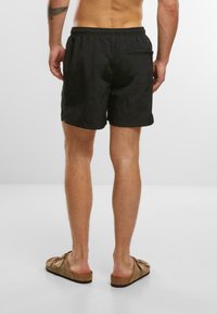 Zwarte zwembroek met een elastische tailleband, achterzak met rits en een gladde textuur. Bruine sandalen met brede banden en platte zolen.