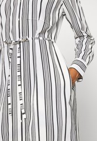 Abito button-up a righe bianche e nere con maniche lunghe, una cintura in vita e una tasca sul petto. Tessuto liscio con linee verticali.