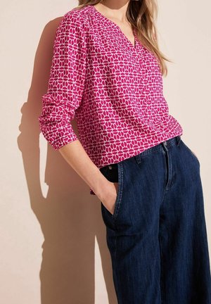 Vrouw draagt een lang mouw roze-wit gebloemde blouse in een high-waist donkerblauwe spijkerbroek met haar handen in haar zakken.