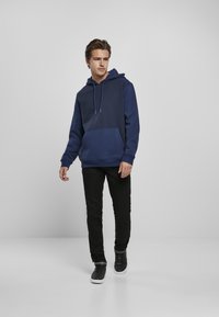 Felpa con cappuccio blu navy con tasca frontale, abbinata a pantaloni neri slim-fit e sneaker neri. Tessuto morbido con design a contrasto.