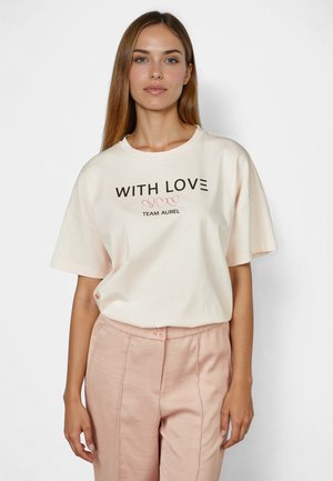 Junge Frau mit langen Haaren, die ein cremefarbenes T-Shirt mit dem Schriftzug "WITH LOVE" und hellrosa Hosen trägt, steht vor einem schlichten weißen Hintergrund.