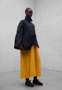 Personne portant une veste sombre et une large jupe jaune, tenant un grand sac cabas noir, debout devant un mur gris clair uni.