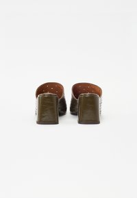 Mules en cuir vert texturé avec un petit talon bloc et un dos ouvert. L'intérieur présente une doublure marron avec des perforations pour la respirabilité.