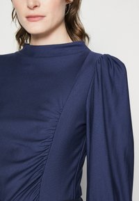 Navy blue dress with a high neck, featuring gathered fabric on one side and puffed sleeves. Smooth texture with a fitted silhouette.