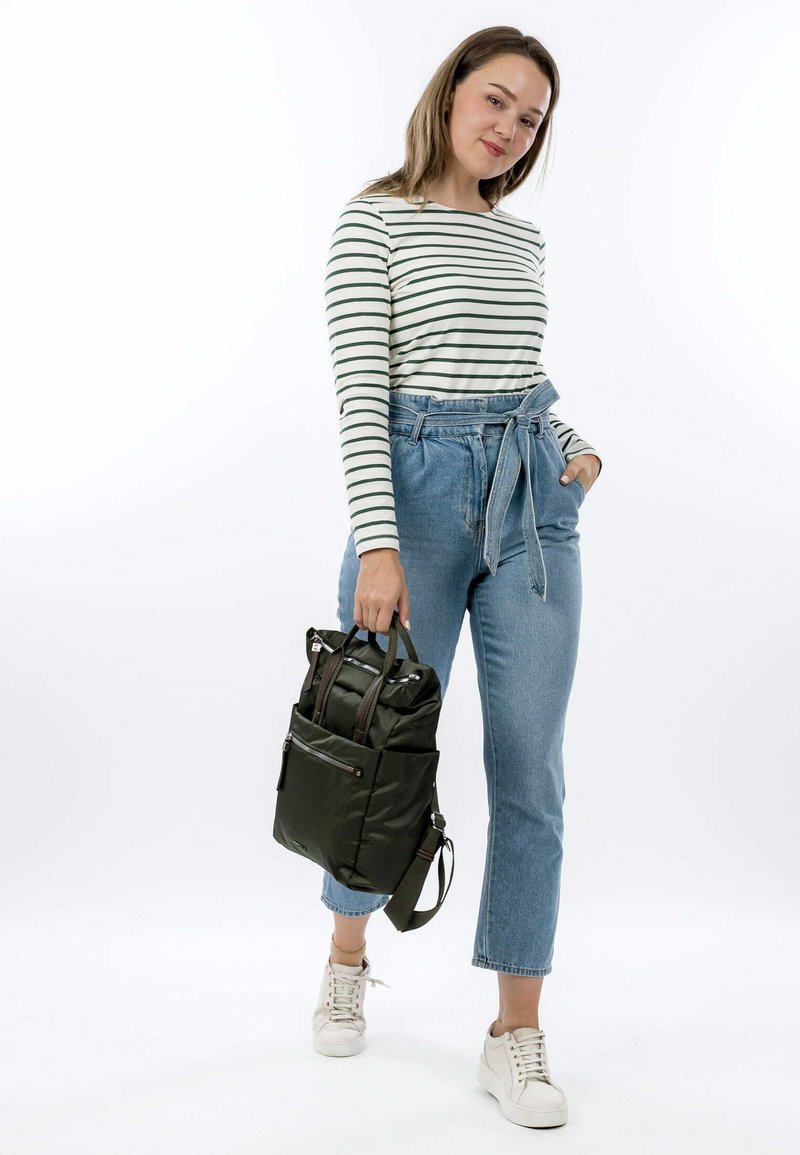 Camicia a righe verdi a maniche lunghe, jeans chiari a vita alta, e sneakers bianche. Porta uno zaino verde scuro con più tasche.