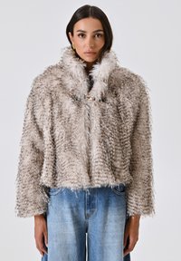 Giacca cropped in pelliccia sintetica di un beige chiaro con macchie più scure, un colletto alto e chiusura a gancio. Indossata sopra jeans a gamba larga blu.