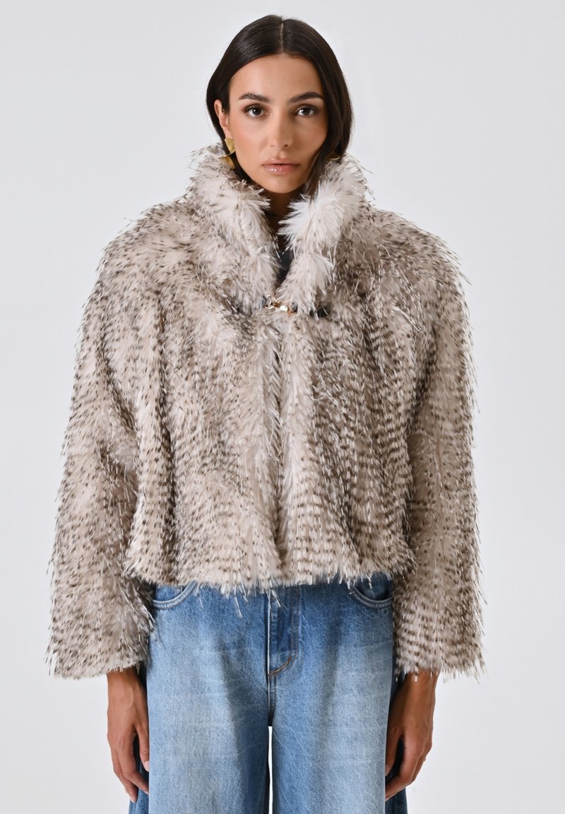 Giacca cropped in pelliccia sintetica di un beige chiaro con macchie più scure, un colletto alto e chiusura a gancio. Indossata sopra jeans a gamba larga blu.