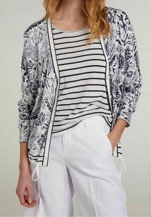 Donna che indossa pantaloni bianchi con tasche, una maglietta a righe bianco e nero e un cardigan bianco con motivi floreali neri.