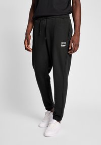 Sorte joggingbukser med snoretræk i taljen, sidelommer og elastiske manchetter. Har hvidt logo på venstre lår. Blød stoftekstur.