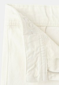 Posnetek v bližini belega džinsa, ki prikazuje nastavljiv pas z gumbom in zadrgo.