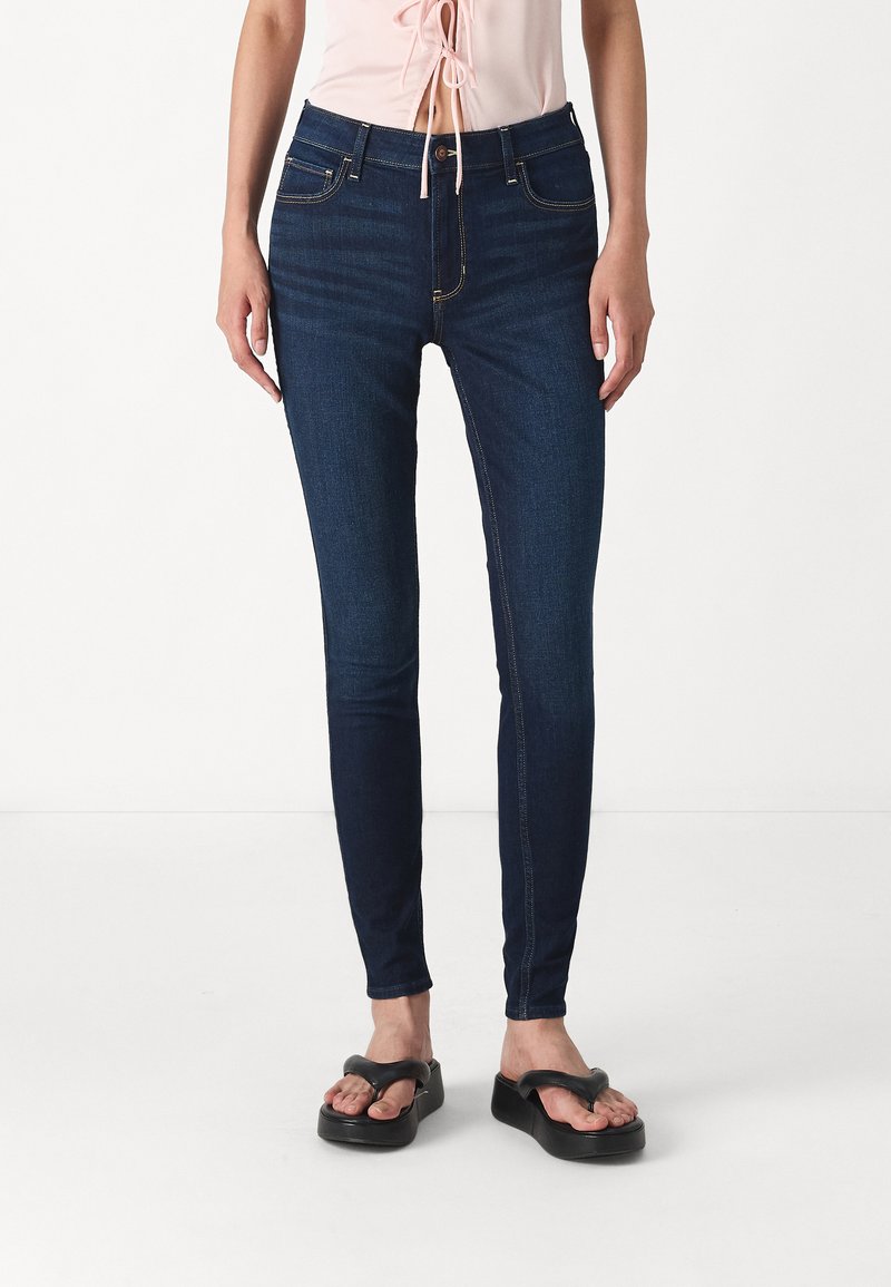 Jeans ajustados de color azul oscuro con una textura suave, diseño de cinco bolsillos y costuras en contraste. Combinados con sandalias negras de tipo chancla.