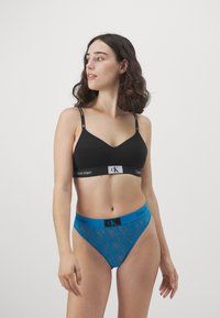 Fekete, varrás nélküli melltartó V kivágással és állítható pántokkal, kék mintás bikini alsóval, logóval ellátott derékkal párosítva.
