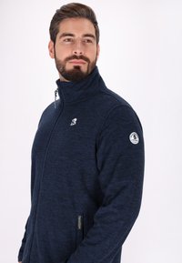 Tengerészeti fleece, cipzáros dzseki magas gallérral, texturált felülettel, hímzett logóval a mellkasán, és oldalsó zsebekkel.