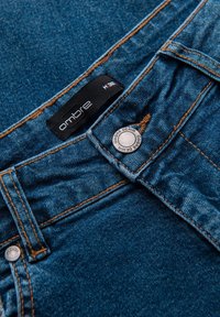 Pantalon en denim bleu foncé avec des surpiqûres orange, doté d'un bouton métallique et d'une étiquette noire portant l'inscription "ombre" près de la ceinture.