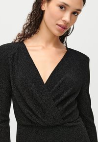 Μαύρο υφαντό φόρεμα με μακριά μανίκια και βαθύ V-ντεκολτέ. Το υλικό έχει μεταλλική λάμψη, με πτυχώσεις στη μέση.