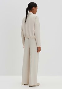 Hellbeiger Overall mit lässigem Schnitt, ausgestattet mit Kragen, elastischem Bund und weitem Bein. Glattes, weiches Material mit minimalen Nähten.