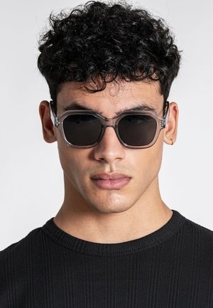 Ung mand med krøllet mørkt hår iført mørke solbriller med gennemsigtigt stel, en lille guldring i øret og en sort ribbet trøje.