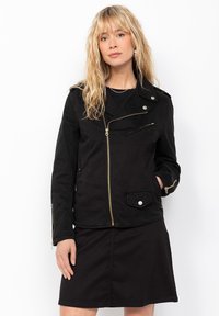 Giacca nera in tessuto con zip frontale, tasche laterali e dettagli metallici; abbinata a una gonna nera.