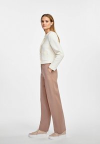 Biała, teksturowana kurtka z postrzępionymi brzegami, jasnoróżowe dopasowane spodnie i beżowe sneakersy. Kurtka posiada kieszeń z przodu oraz zapięcie na guziki.