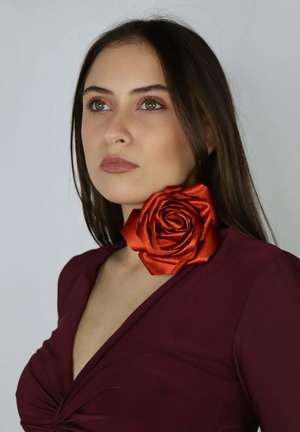Kobieta o długich ciemnych włosach nosi duży czerwony aksamitny kwiat różany na szyi oraz głęboko bordową bluzkę z dekoltem w serek, spoglądając nieco do góry.