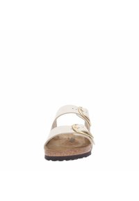 White leather sandal with two adjustable straps, gold hardware, cork footbed, and black rubber outsole, featuring a flat silhouette.