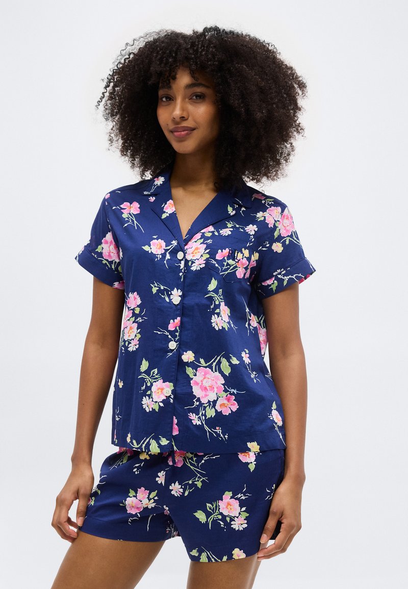 Γυναίκα που φοράει μπλε-σκούρο μπλουζάκι με κοντά μανίκια και κουμπιά μπροστά μαζί με ταιριαστό σορτς με ροζ και λευκή floral εκτύπωση, στέκεται μπροστά σε λευκό φόντο.