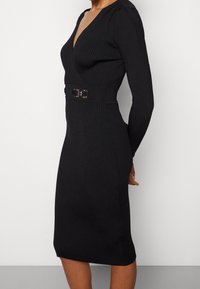 Schwarzes geripptes Strickkleid mit langen Ärmeln, tiefem V-Ausschnitt, taillierter Passform und goldfarbener Akzent-Gürteldetails. endet knapp über dem Knie.