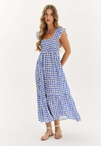 Modrá a biela károvaná maxi šaty so smockovaným živôtikom, volánovými krátkymi rukávmi a vrstveným lemom. Obsahuje malé kvetinové vzory po celom povrchu.