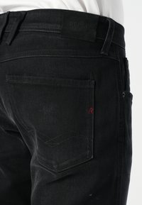 Mustmustest denim püksid, millel on nähtav tagatasku punaste õmblustega, reljeefne logo silt ja peened õmblusdetailid taskutel.