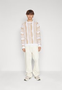 Långärmad vit topp med bruna mönstrade designer, parat med lösa krämfärgade byxor och grå sneakers, stående mot en enfärgad bakgrund.