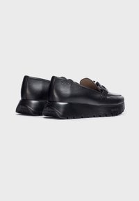 Scarpe slip-on in pelle nera con suola in gomma spessa, caratterizzate da una finitura liscia e un design curvo, accentuate da un piccolo dettaglio decorativo.