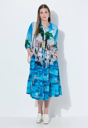 Frau steht und trägt ein langes blaues Kleid mit buntem Aufdruck eines Küstenortes, weiße Plateauschuhe und trägt eine hellfarbige Umhängetasche.