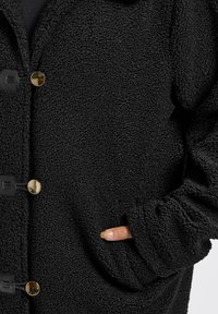 Svart texturerad kappa med en mjuk, lurvig yta. Har guldknappar och sidofickor, med en avslappnad passform och uppvikta ärmar.