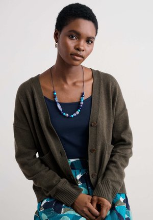 Olive green cardigan with a soft texture, featuring ribbed cuffs, front pockets, and brown buttons over a navy top and patterned skirt.
